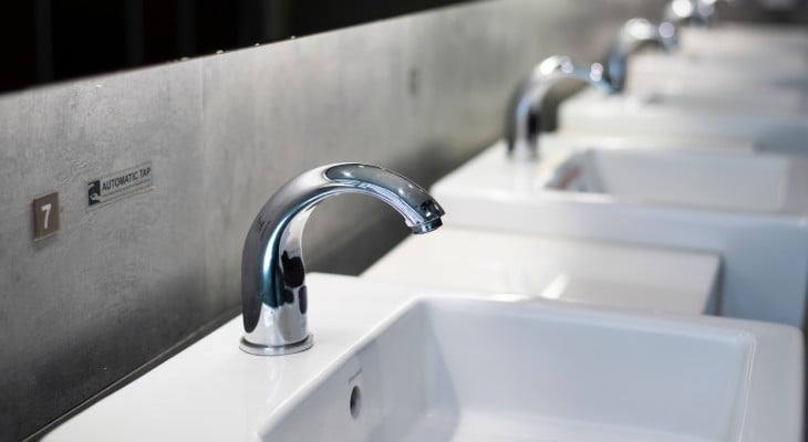 A row of automatic faucets attached to clean white sinks lined up against the wall of a modern commercial bathroom.