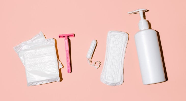 Two sanitary napkins, a tampon, an open pad, a bottle of lotion, and a razor lined up against a pink background.