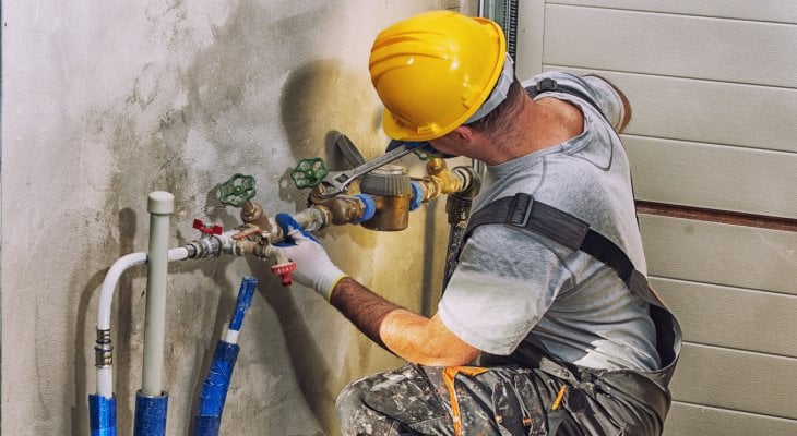 A plumber wearing a hard hat and protective gloves is bent down using a wrench to tighten a valve on a pipe.